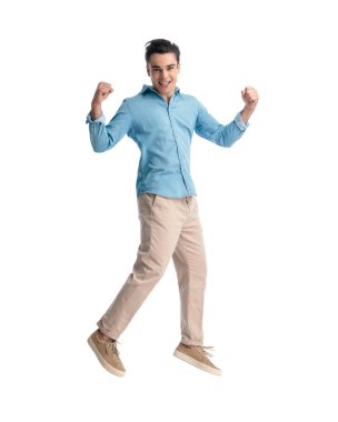 Mavi kot gömlekli heyecanlı genç adam, beyaz arka planda kazanmak için yumruklarını kaldırıp havaya zıplıyor.