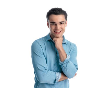 Mavi kotlu gömlekli mutlu bir adam kollarını katlıyor, çenesini yumrukluyor ve beyaz arka planda poz verirken gülümsüyor.