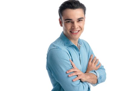 Mavi kot gömlekli, kollarını kavuşturup beyaz arka planda dururken gülümseyen kendine güvenen genç adamın yan görüntüsü.