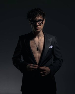 Gözlüklü, siyah ceketli, göğsünü gösteren ve karanlık arka planda poz veren seksi, zarif bir adam.