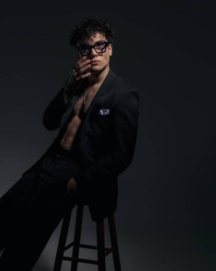 Siyah ceketli düşünceli seksi adam kaslarını gösteriyor, yüzüne dokunuyor, yukarı bakıyor ve karanlık arka planda otururken düşünüyor.