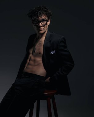 Siyah ceketli çekici bir adam. Kaslarını ve göğsünü gösteriyor. Gözünü kaçırıyor ve arka planda tahta bir sandalyede oturuyor.