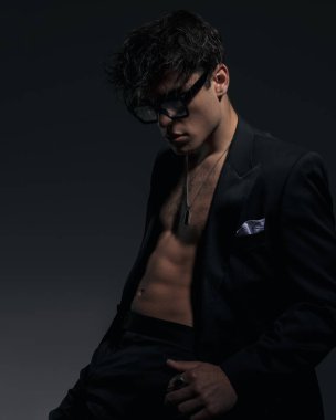 Siyah gözlüklü, baştan çıkarıcı genç adam aşağı bakarken ve arka planda otururken kaslarını gösteriyor.