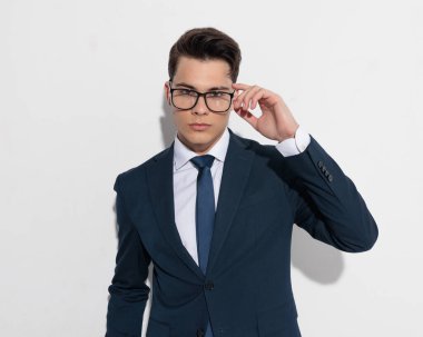 Lacivert takımlı, kendine güvenen genç işadamı gözlüklerini ayarlıyor ve gri arka planda havalı bir poz veriyor.