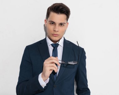 Lacivert takımlı seksi, zarif adam gözlüklerini çıkarıyor ve gri arka planda kendine güvenen bir şekilde kameraya bakıyor.