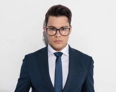 Gözlüklü, lacivert takımlı, kravatlı, gri arkaplan önünde poz veren zarif bir genç.