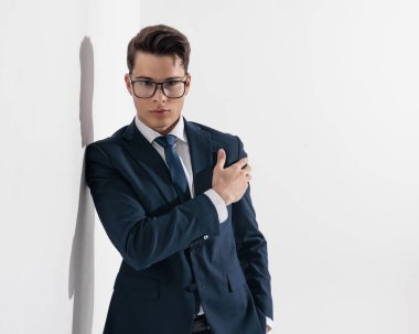 Gri arkaplan önünde koluna dokunurken duvarda gözlük takan lacivert takımlı zarif genç adam.