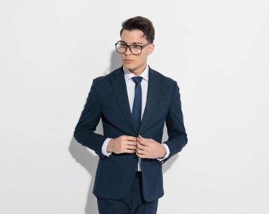 Gözlüklü zarif genç işadamı gri arkaplan önünde lacivert takım elbise giyip kapatıyor.