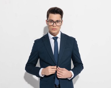 Gözlüklü yakışıklı genç işadamının portresi. Gri arka planın önünde lacivert takım elbise ilikliyor.