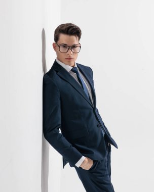 Lacivert takım elbiseli, duvarda gözlüklü ve gri arka planda elleri ceplerinde poz veren seksi, zarif bir genç.