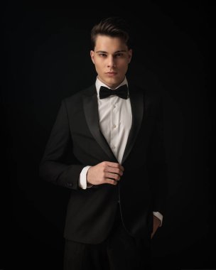 Kendine güvenen genç damat zarif siyah smokinini ayarlıyor ve siyah arka planda el ele poz verirken ileriye bakıyor.