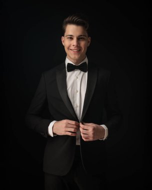 Siyah smokinini ilikleyip gülümseyen genç damat siyah arka planda durup poz verirken.