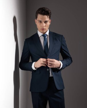 Kendine güvenen genç adam gri duvarın önünde beklerken lacivert takımının düğmelerini ilikliyor.