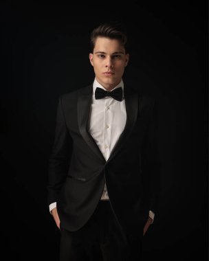 Siyah zarif smokinli çekici bir genç ceplerinde el ele tutuşup siyah arka planda poz veriyor.