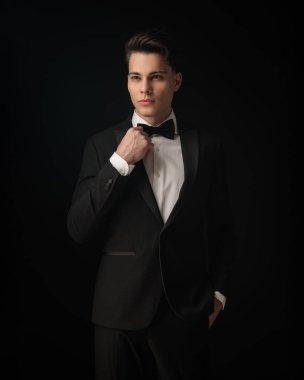 Siyah smokinli yakışıklı damat gözlerini kaçırıyor, siyah smokini ayarlıyor ve siyah arka planda el ele poz veriyor.