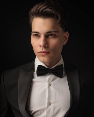 Smokinli, papyonlu, siyah arka planda poz veren çekici bir genç.