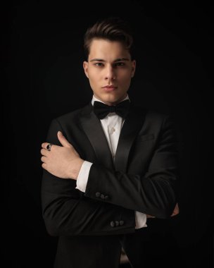 Siyah smokinli, papyonlu, kollarını kavuşturup siyah arka planda poz veren kendine güvenen bir damat.