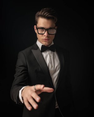 Siyah smokinli yakışıklı genç adam. Gözlüklü, el ele tutuşmuş ve siyah arka planda poz veriyor.