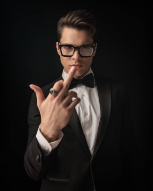 Siyah smokinli zarif bir genç. Gözlüklü. Orta parmağını siyah arka plan önünde saldırgan bir hareket olarak gösteriyor.