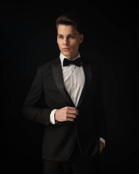 confident elegant man adjusting black tuxedo and looking away while posing with hand in pocket on black background