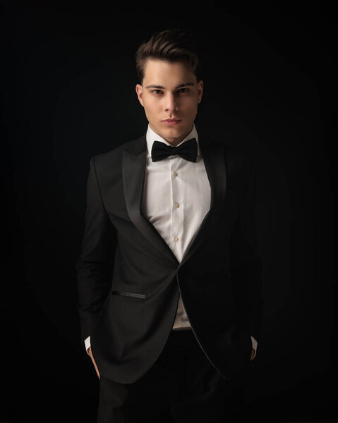 young elegant groom in black tuxedo holding hands in pockets and standing in front of black background
