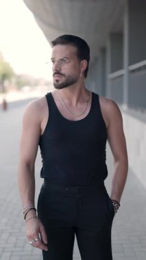 Gözlüklü, siyah tişörtlü havalı adam yan bakıyor, el ele tutuşuyor, avuçlarını ovuyor, güvenli bir şekilde dışarıda yürüyor.