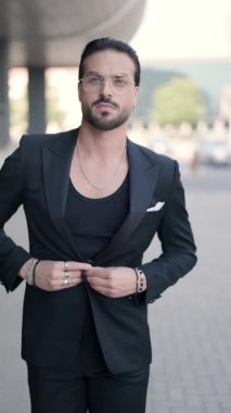 Gözlüklü, siyah takım elbiseli, yürüyen, yana bakan, kendine güvenen, el ele tutuşan, avuçlarını ovuşturan şık havalı bir adam.