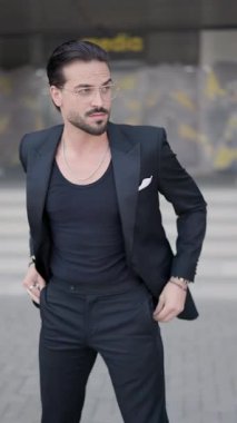 Tıraş olmamış çekici bir iş adamı, takım elbise ceketini açıyor, elini cebine sokuyor ve yürüyor, etrafa bakıyor ve ceketini ilikliyor.