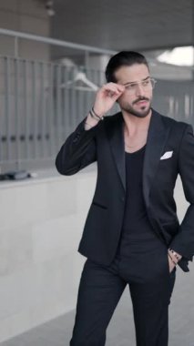 Taraf tutan seksi iş adamının dikey videosu. Gözlük ve ceketi tamir ediyor, elleri ceplerinde yürüyor.