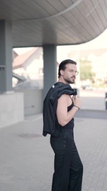 Seksi sakallı adam ceketini omuzunda tutuyor, hareket ediyor, gözlükleri ayarlıyor, ileriye bakıyor ve baştan çıkarıcı bir şekilde gülümsüyor.