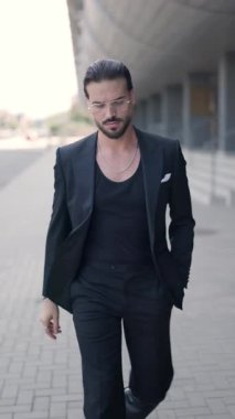 Siyah takım elbiseli zarif genç adam el ele hareket ediyor, etrafa bakıyor ve gözlükleri ayarlıyor.