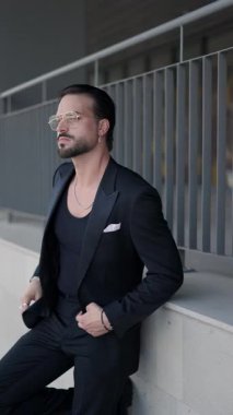 Gözlüklü, siyah takım elbiseli seksi genç işadamı, yan tarafa bakıyor, dirseğini duvara dayayıp gülüyor.