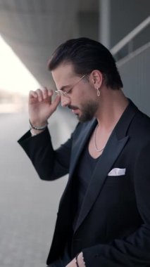 Zarif bir adamın gözlük takması, siyah ceketi iliklemesi, çeneye dokunması, kollarını kavuşturması ve poz vermesi.