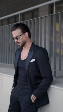 Havalı güneş gözlüklü, cebinde el ele tutuşan, siyah takımı kapatan ve dışarıdayken gülümseyen seksi ve zarif bir adam.