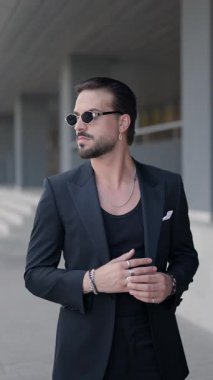 Çekici moda adamı, havalı güneş gözlüklerini takıyor ve yan tarafa bakıyor, kendine güveniyor ve gülümsüyor, kamera yakınlaşırken.