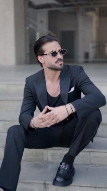 Kendine güvenen genç bir adam, dizlerini dizlerinin üstüne çökerek ve merdivenlerde oturarak güneş gözlüğünü takmaya çalışıyor.