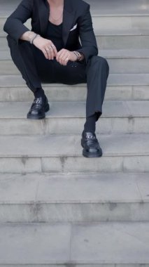 Siyah takım elbiseli, güneş gözlüklü, dizlerinin üstünde dirseklerini tutan ceketini ayarlayan ve şehir merdivenlerinde oturan seksi bir genç.