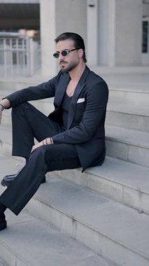 Gözlüklü, dizinde dirsek tutan, öne ve yana bakan ve şehir merdivenlerinde otururken ceketini ayarlayan zarif bir adamın yan görüntüsü.