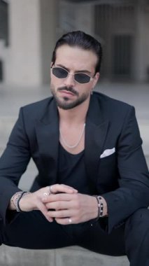 Güneş gözlüklü, siyah takım elbiseli, dirseği merdivenlerde uzanmış, yan tarafa bakan seksi bir adamın dikey videosu.