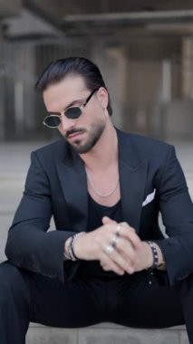 Siyah takım elbiseli, güneş gözlüklü zarif genç adam kollarını yan yana tutuyor, dirseğiyle merdivenlerde oturuyor ve güneş gözlüğünü tamir ediyor.