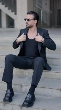 Oturmuş, güneş gözlüklü, siyah takım elbiseli, dirseği dizinde oturan ve kendine güvenen bir şekilde poz veren zarif bir iş adamı.