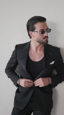 Siyah takım elbiseli, elini cebine sokup gri arkaplanın önünde duran seksi, zarif iş adamının dikey videosu.