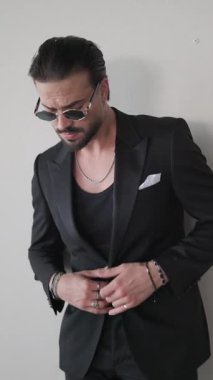 Havalı, kibirli, ceketinin düğmelerini çözen ve ellerini arka planda gri ceplere koyarak yan tarafına bakan adam.