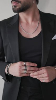 Siyah takım elbiseli, gümüş yüzüklü, elleri cebe sokan ve gri arka planda güvenle poz veren seksi bir genci gösteren videoyu kapat.