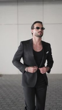Güneş gözlüklü kendine güvenen moda adamı siyah ceketini kapatıyor ve elleri ceplerinde yürüyor, yana bakıyor ve gözlükleri tamir ediyor.