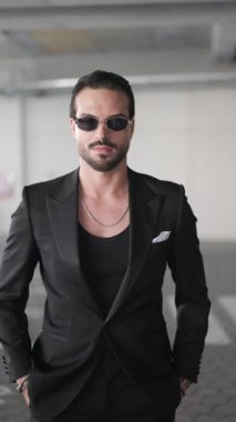 Siyah takım elbiseli çekici, sakallı, güneş gözlüklü, el ele tutuşan ve şehvetli bir şekilde gülümseyen bir adam.