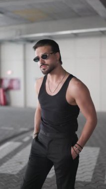 Siyah tişörtlü seksi adam kollarını kavuşturuyor, yan yana bakıyor ve dışarıda dururken güneş gözlüğünü ayarlıyor.