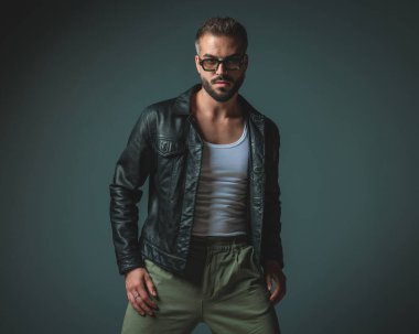 Sokak modasına uygun, güneş gözlüklü genç adam gri arka planda siyah deri ceket giyerken havalı poz veriyor.