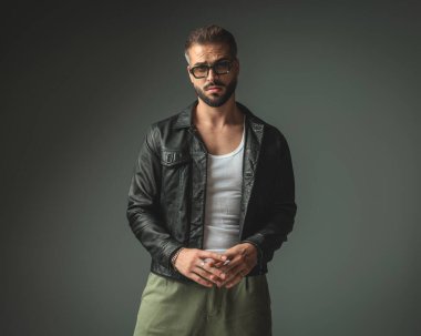 Güneş gözlüklü, siyah deri ceketli, ellerine dokunan ve gri arka planda duran çekici bir adam.