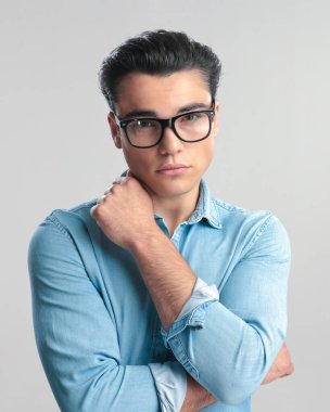 Kollarını kavuşturan gözlüklü, mavi kot pantolonlu seksi genç adam gri arka planda poz verirken boynuna dokunuyor.